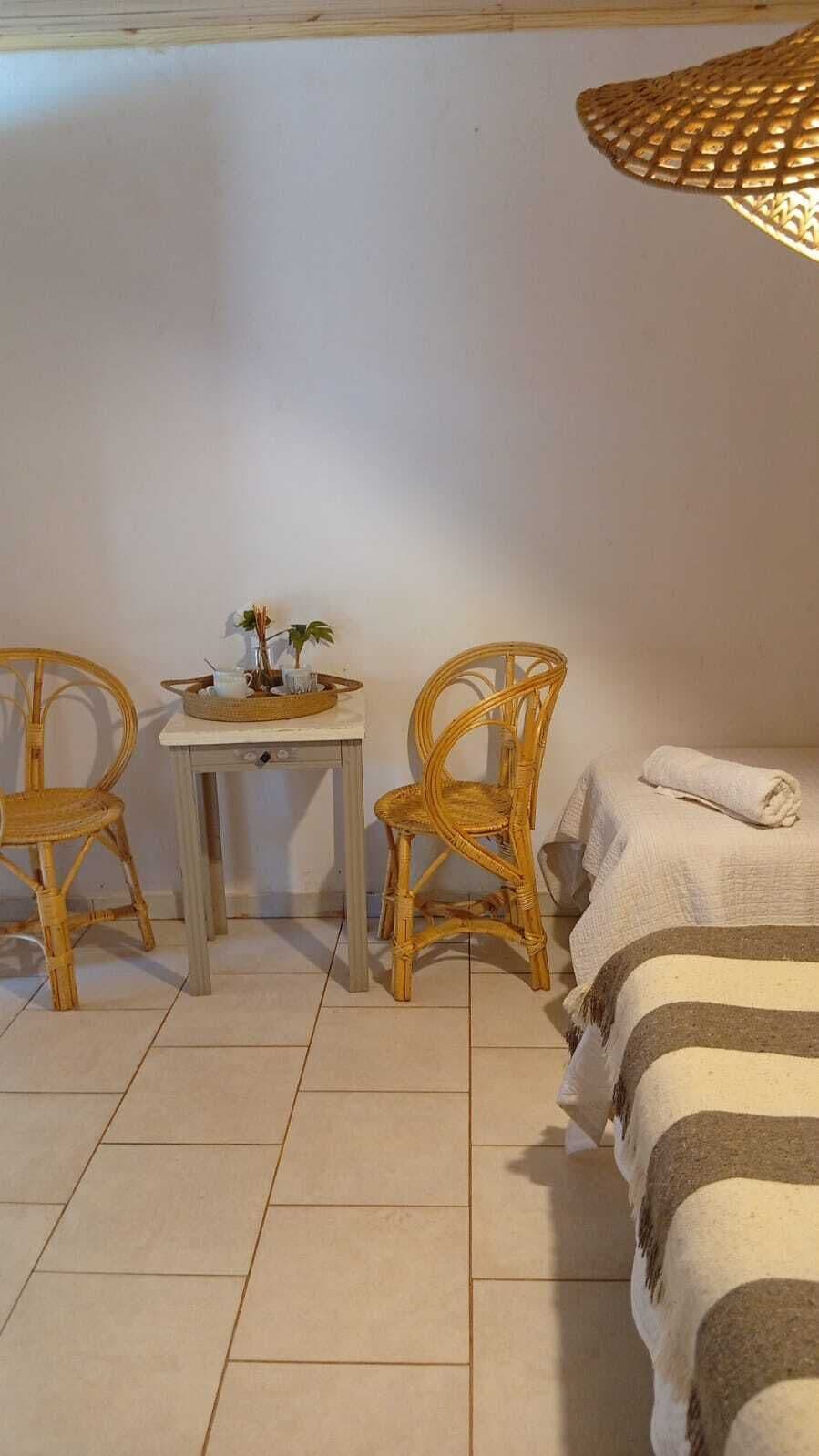 A room with a bed, two wicker chairs, a small table with a tray, and a woven light fixture.