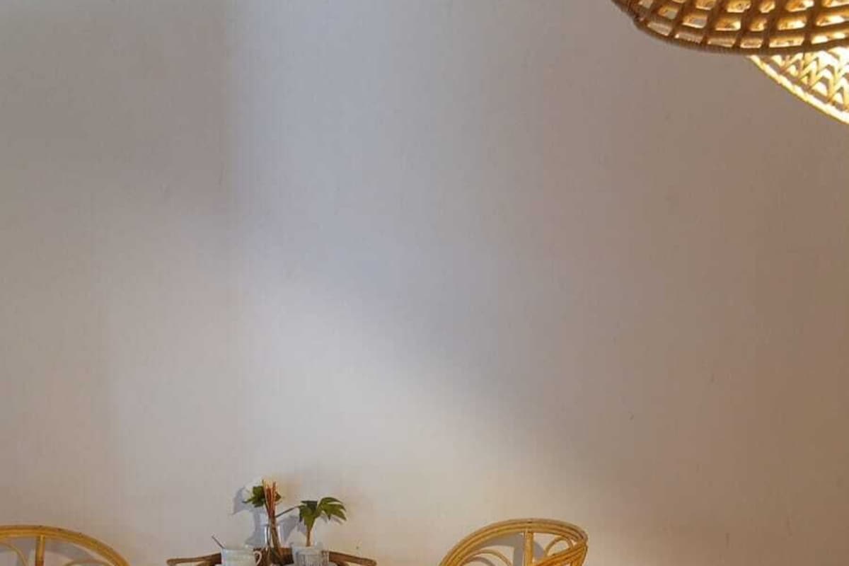 Una habitación con una cama, dos sillas de mimbre, una mesita con bandeja y una lámpara de fibra trenzada.