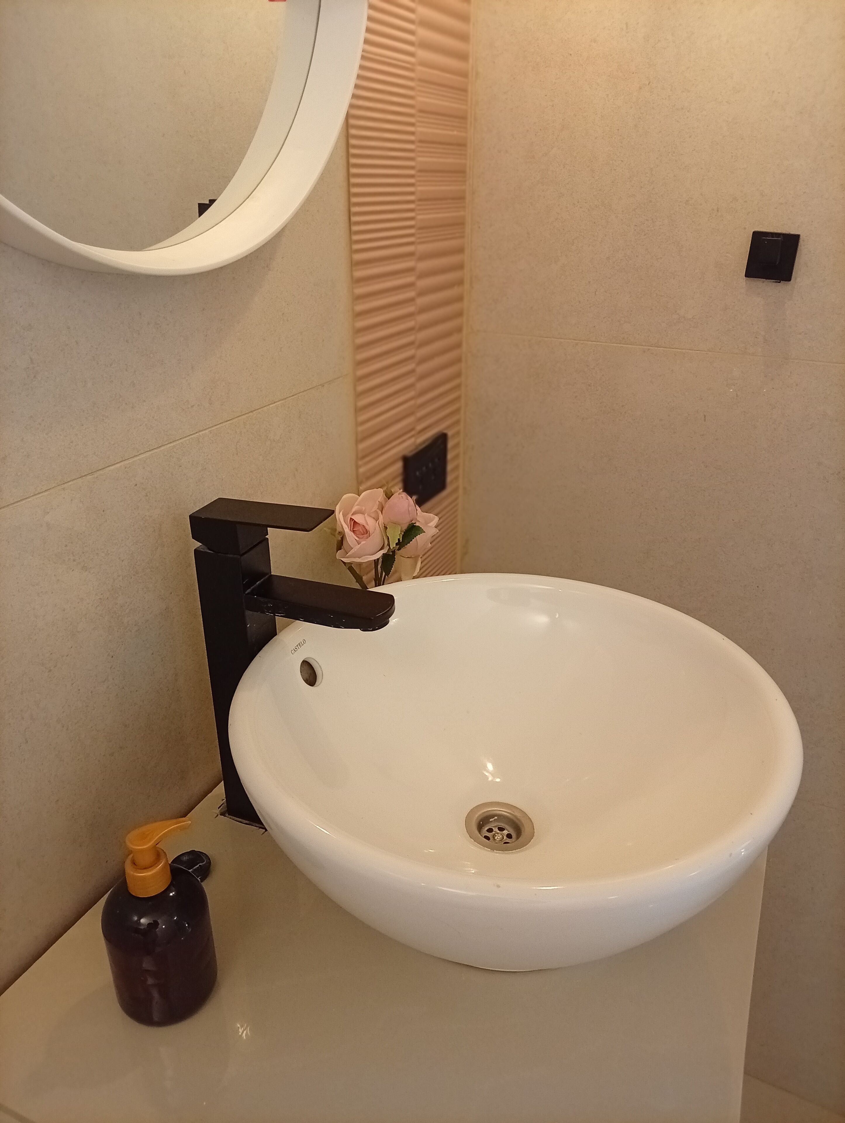Casa de banho com lavatório branco, torneira preta, espelho e um pequeno vaso com flores.