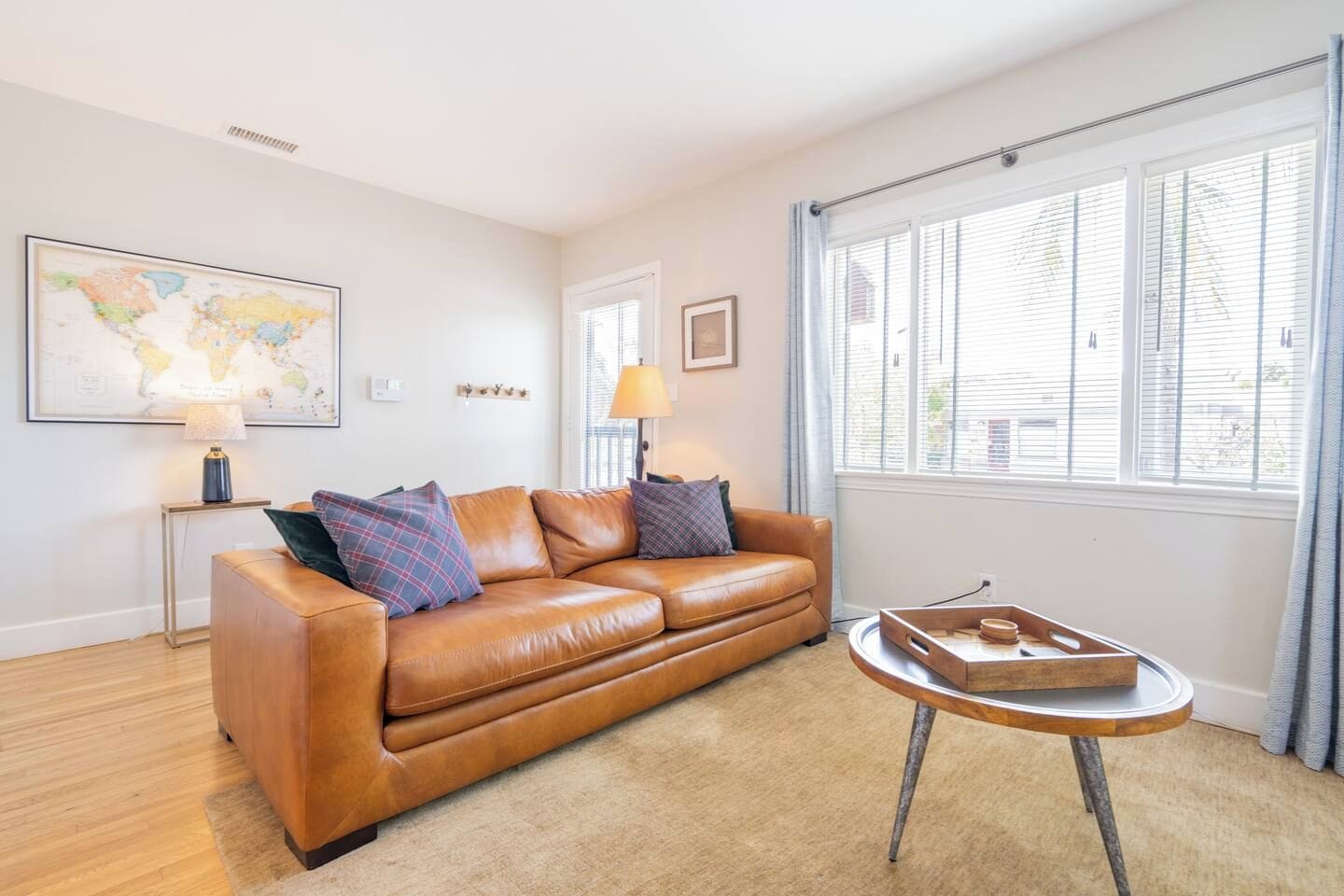 A furnished living room with a leather sofa, a round coffee table, a world map on the wall, and a window with blinds.