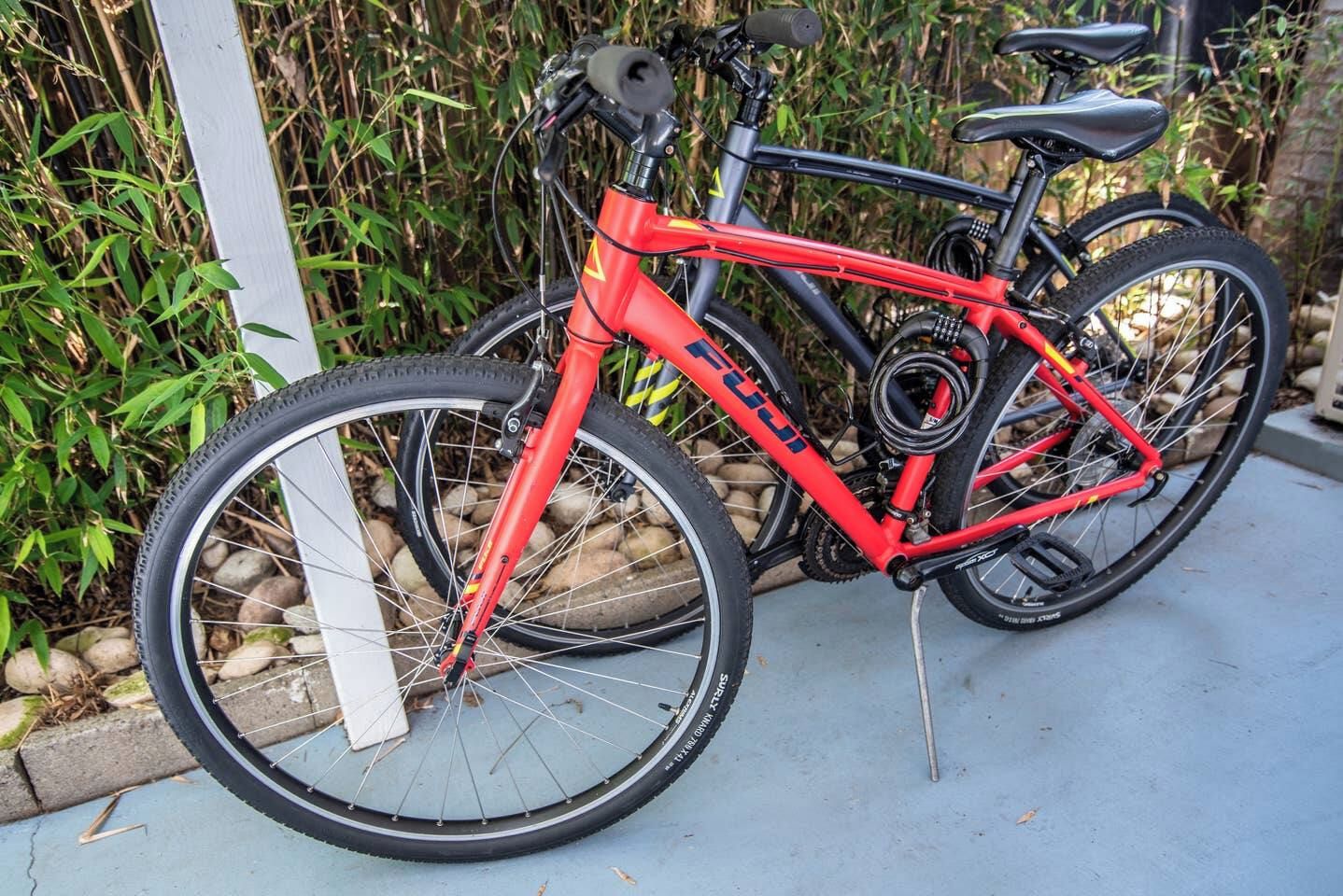 Uma bicicleta vermelha com selim e guidão pretos, estacionada sobre uma superfície de concreto, com um poste branco e vegetação verde ao fundo.