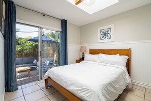 Ein Schlafzimmer mit einem Bett, einem Nachttisch, einer Lampe, einem Fenster mit Vorhängen und Blick auf einen Außenbereich mit Sofa und Sessel.