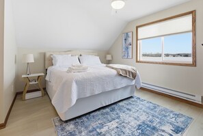 A bedroom with a bed, bedside tables, a lamp, a window with a view, and a painting on the wall. - Spacious lakefront retreat with Private Dock, Fire Pit and Sunset Views. (Houghton Lake)