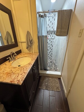 Un baño con lavamanos de mármol, armarios de madera oscura, ducha con pared de azulejos y una pequeña alfombra en el suelo.
