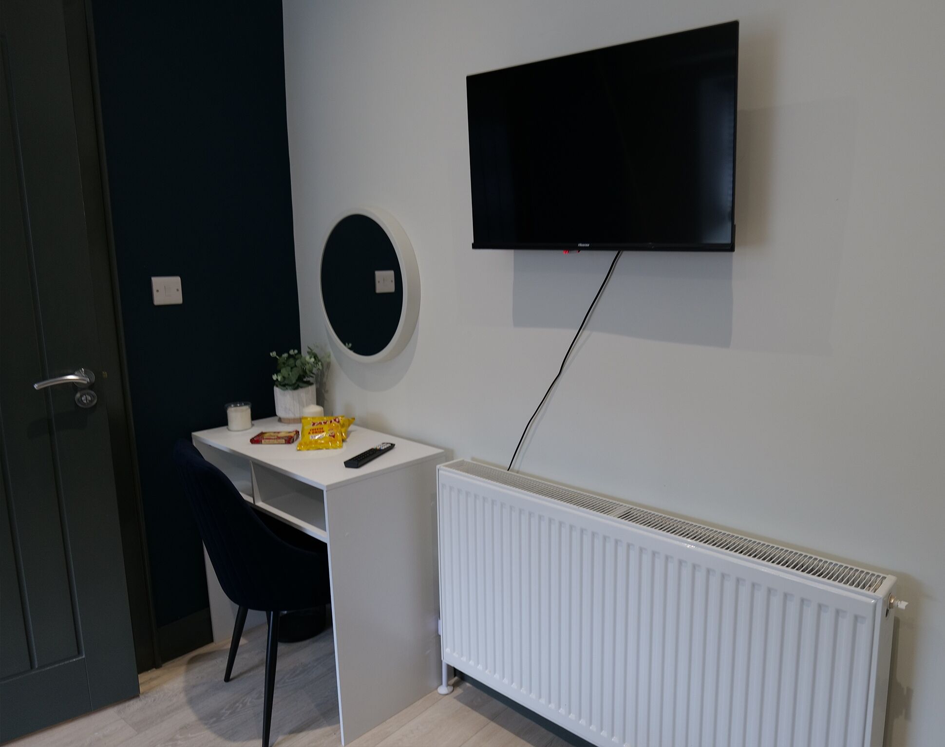 A room with a white desk, a black chair, a mounted TV, a white radiator, and a door.