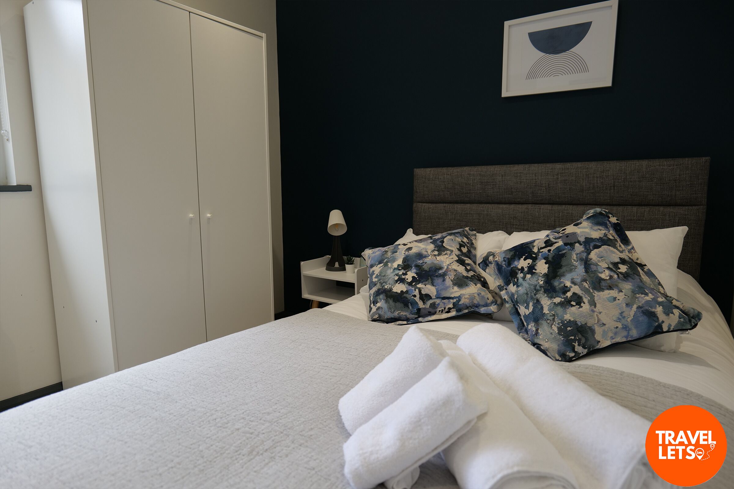 Un lit bien fait, avec du linge de lit blanc et une pile de serviettes pliées, une table de chevet avec une lampe, et un mur sombre orné d’un tableau encadré.