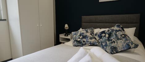 A neatly made bed with white linens and a pair of folded towels, a nightstand with a lamp, and a dark wall with a framed picture.