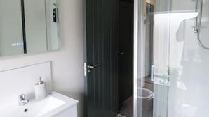 Ein modernes Badezimmer mit einem weißen Waschbecken, einem Spiegel, einer dunklen Tür und einer Duschkabine aus Glas.