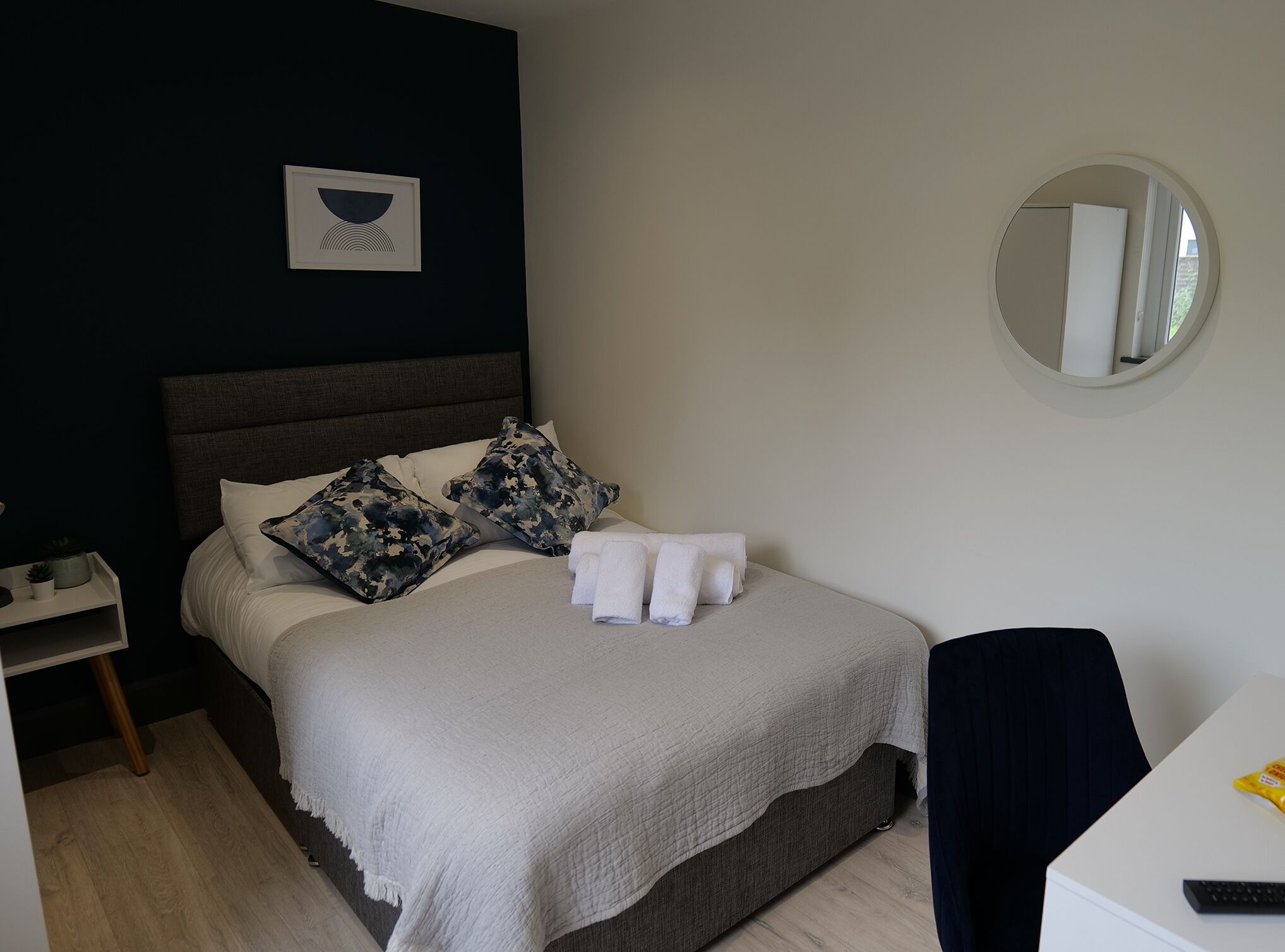A neatly made bed with white linens and two camouflage pillows, a bedside table with a plant, and a round mirror on the wall.