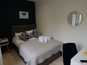 Una cama bien hecha con sábanas blancas y dos almohadas de camuflaje, una mesita de noche con una planta y un espejo redondo en la pared.
