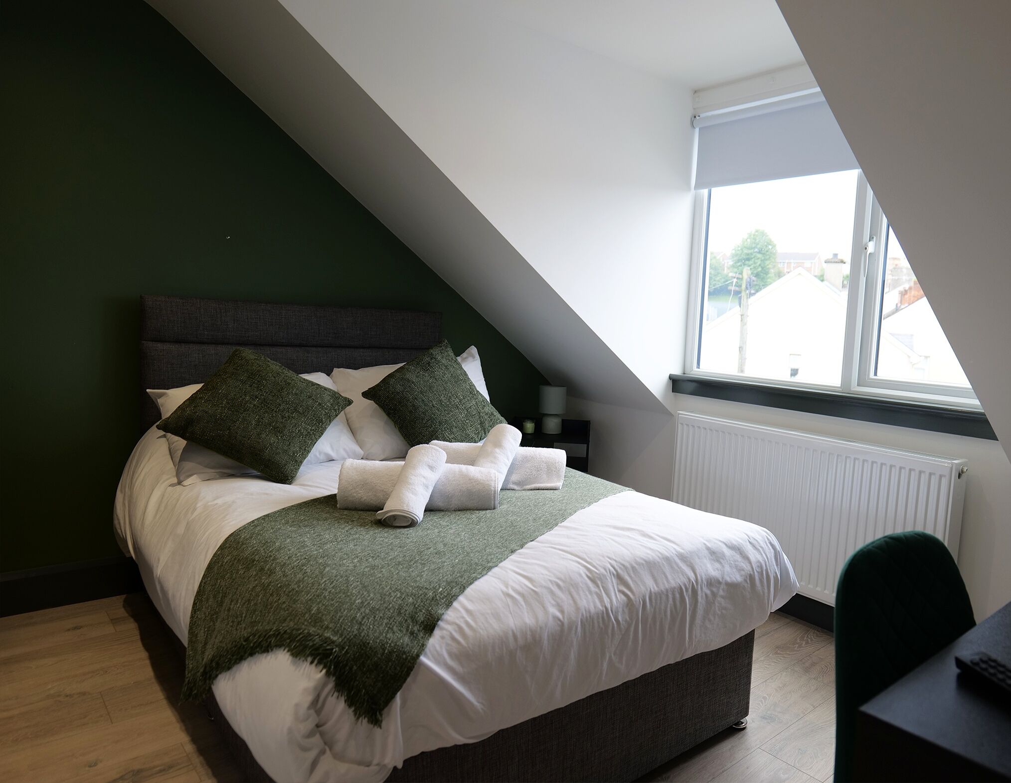Ein Schlafzimmer mit Bett, Kissen, Decke, Heizkörper, Fenster und Schreibtisch.