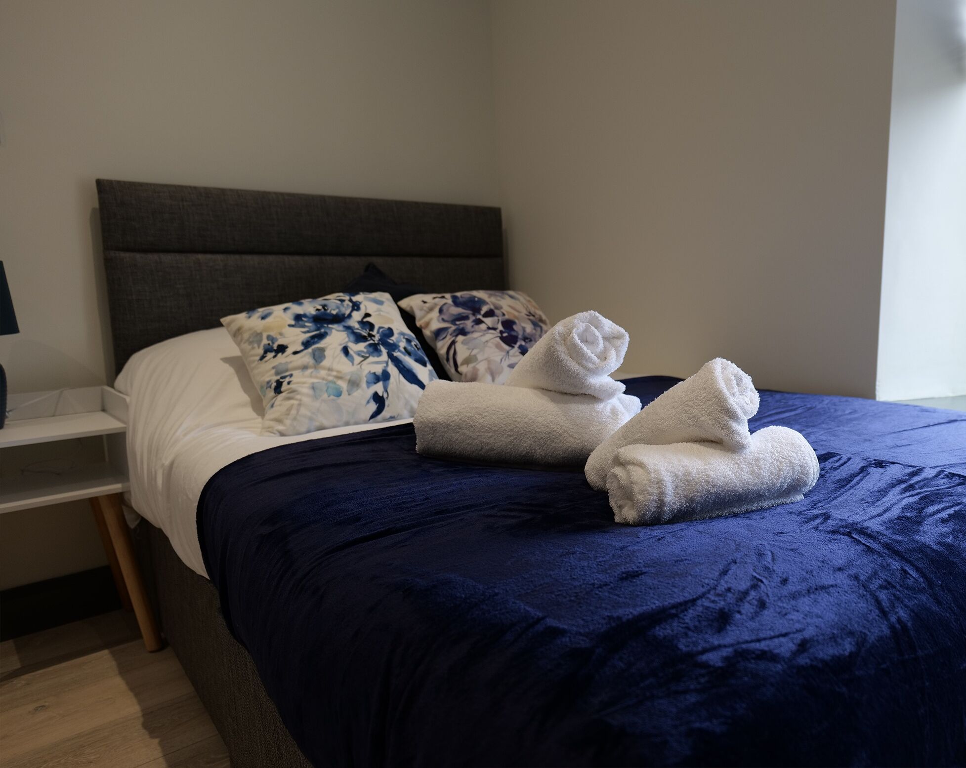Un lit bien fait, recouvert d’une couvre-lit bleu foncé, avec deux oreillers à motifs floraux et deux serviettes blanches pliées.