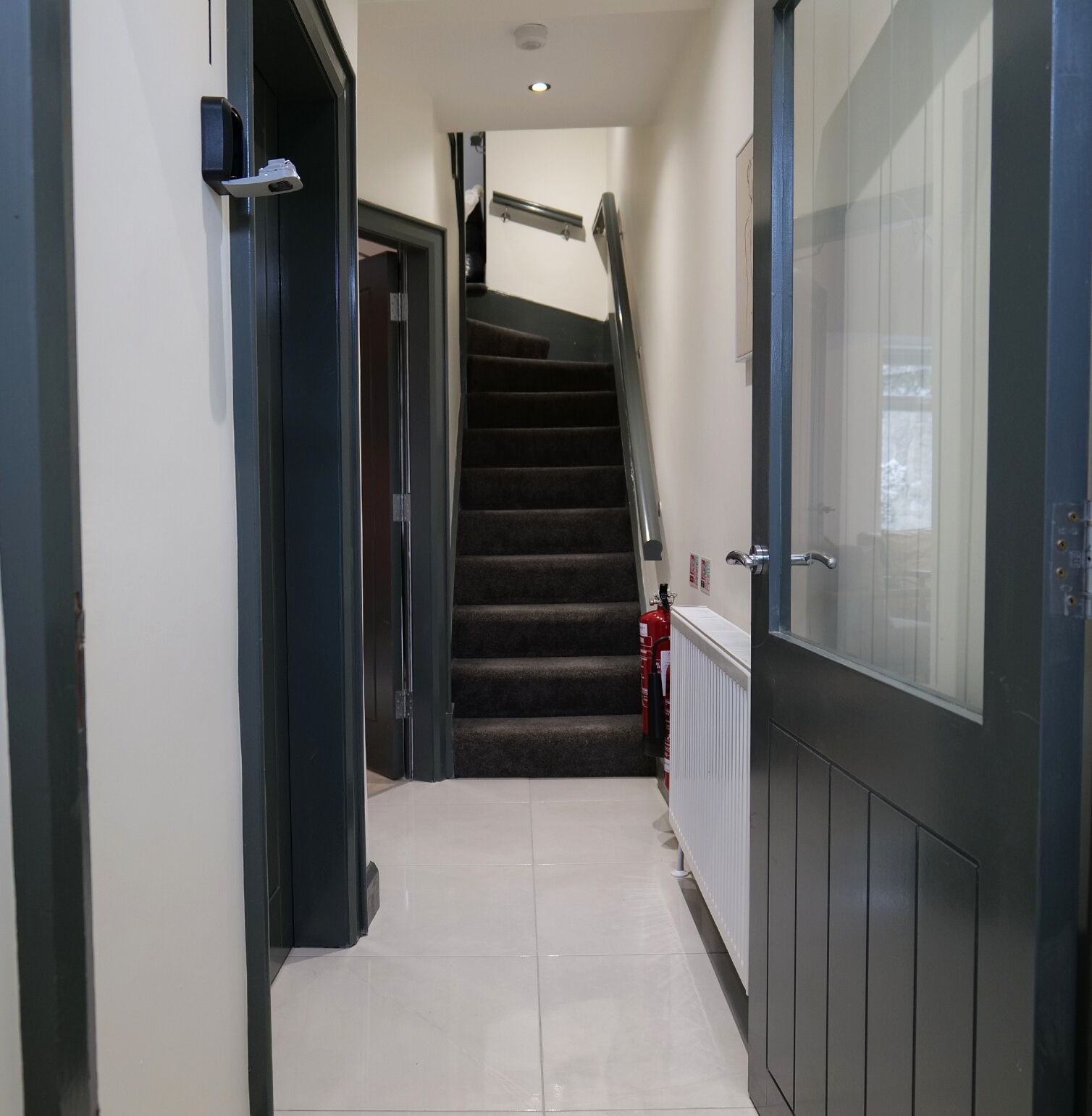 A corridor with a staircase leading to an upper floor, a fire extinguisher, and a door with a handle.