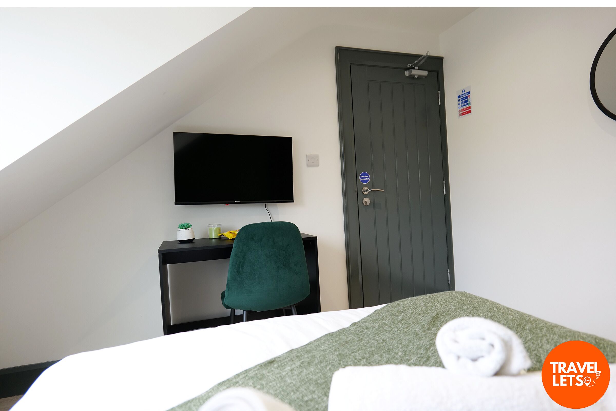 Habitación de hotel con cama, escritorio con televisor, una silla verde y una puerta con letrero.