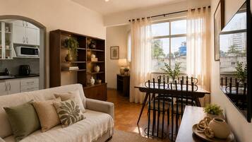 Uma sala de estar aconchegante com sofá, mesa de jantar com cadeiras, cozinha com armários e micro-ondas, e uma grande janela com cortinas.