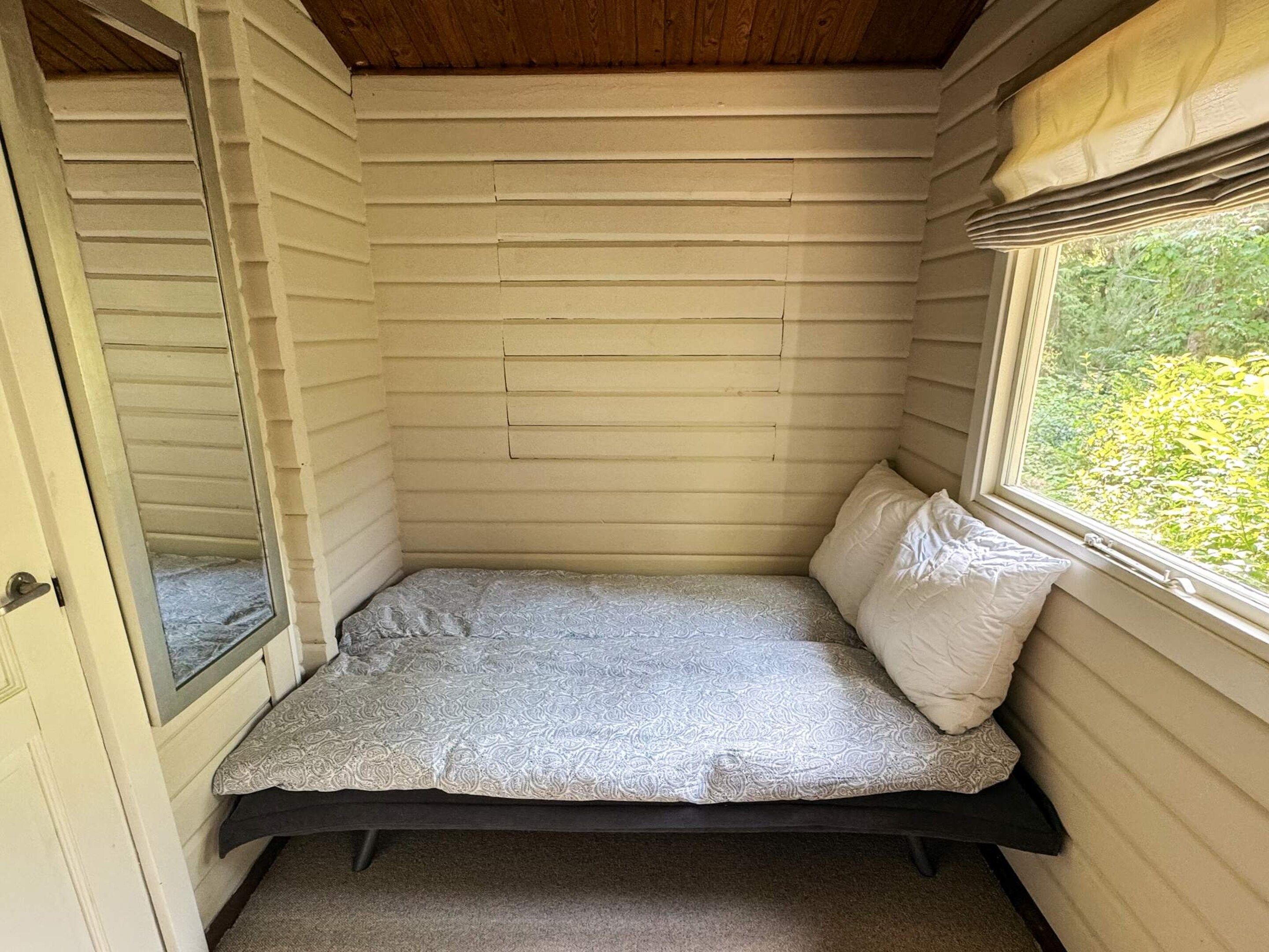Une petite pièce fermée, avec un lit simple, une fenêtre et une porte.
