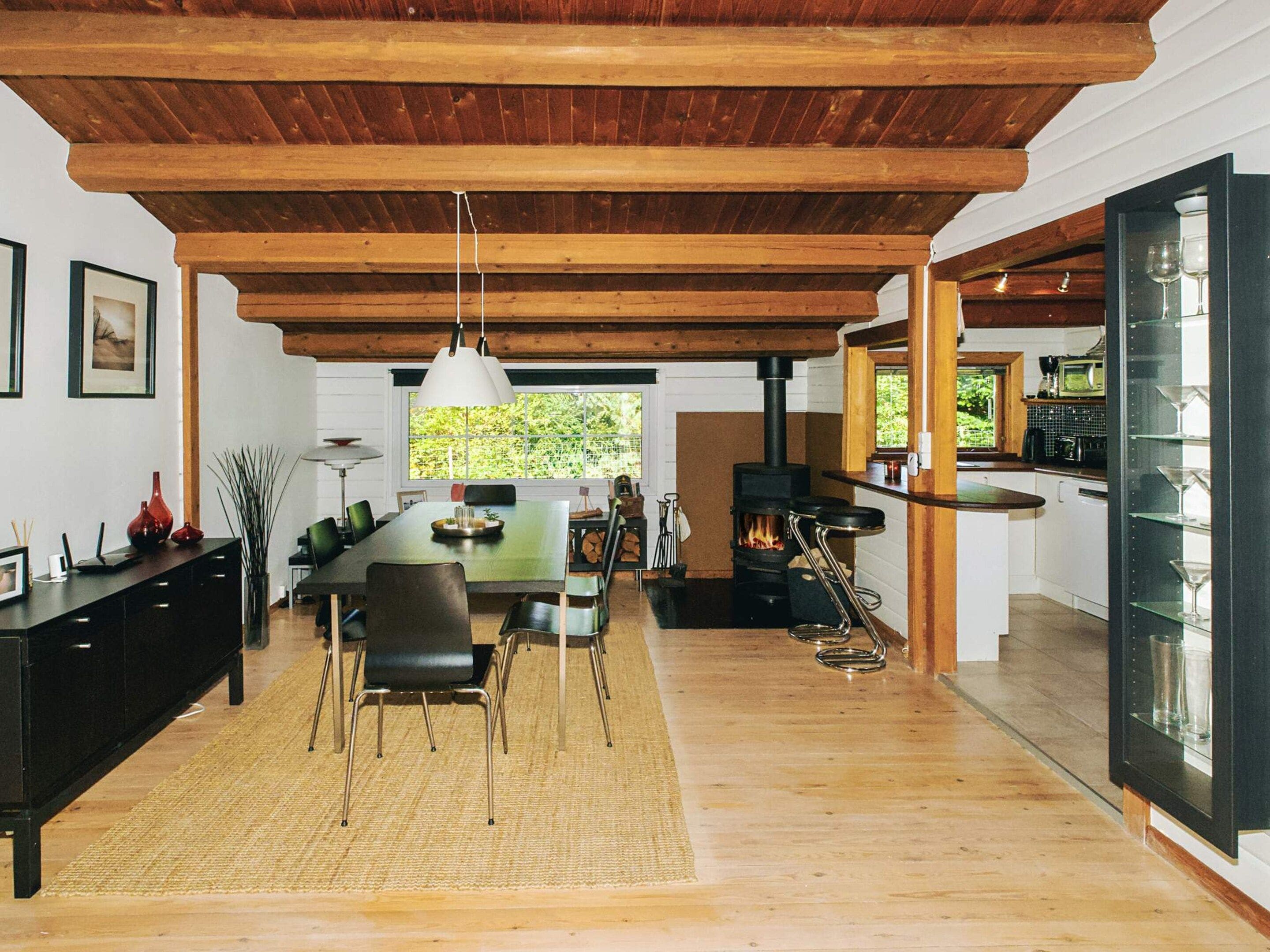 A spacious dining area with a wooden ceiling, a large table, and chairs. A modern kitchen with a stove and glassware is visible to the right.