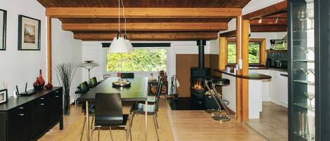 A spacious dining area with a wooden ceiling, a large table, and chairs. A modern kitchen with a stove and glassware is visible to the right.
