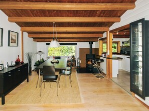 Een ruime eethoek met een houten plafond, een grote tafel en stoelen. Een moderne keuken met een fornuis en glazen is rechts zichtbaar.