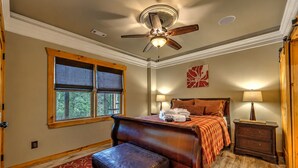 Habitación de hotel con cama, escritorio, silla, mesita de noche, lámpara, ventilador de techo, ventana con persianas y puerta.