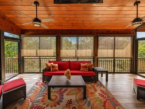 A covered patio with a red sofa, two wicker chairs, a coffee table, and a patterned rug. - Come Vacation at Home - 6.5 Acres, Hot Tub, Game Room, Mins to Blue Ridge (Mineral Bluff)