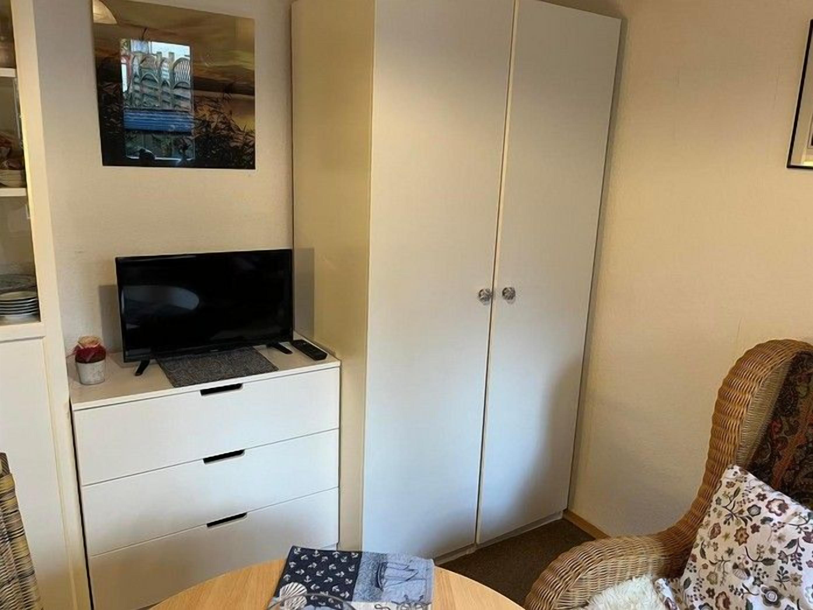 Quarto com televisão em suporte, guarda-roupa, cama com almofada estampada e pequena mesa com um livro.