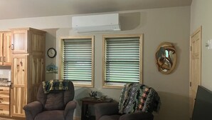 Een woonkamer met twee fauteuils, een bijzettafeltje en een televisie. Er zijn twee ramen met zonwering, een klok en een spiegel aan de muur.