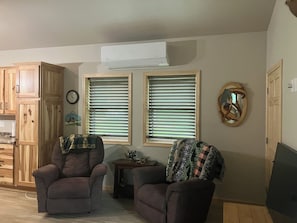 A living room with two armchairs, a small table, and a television. There are two windows with blinds, a clock, and a mirror on the wall. - Enjoyable 2-bedroom cabin with AC in Crivitz near the high Falls flowage (Crivitz)