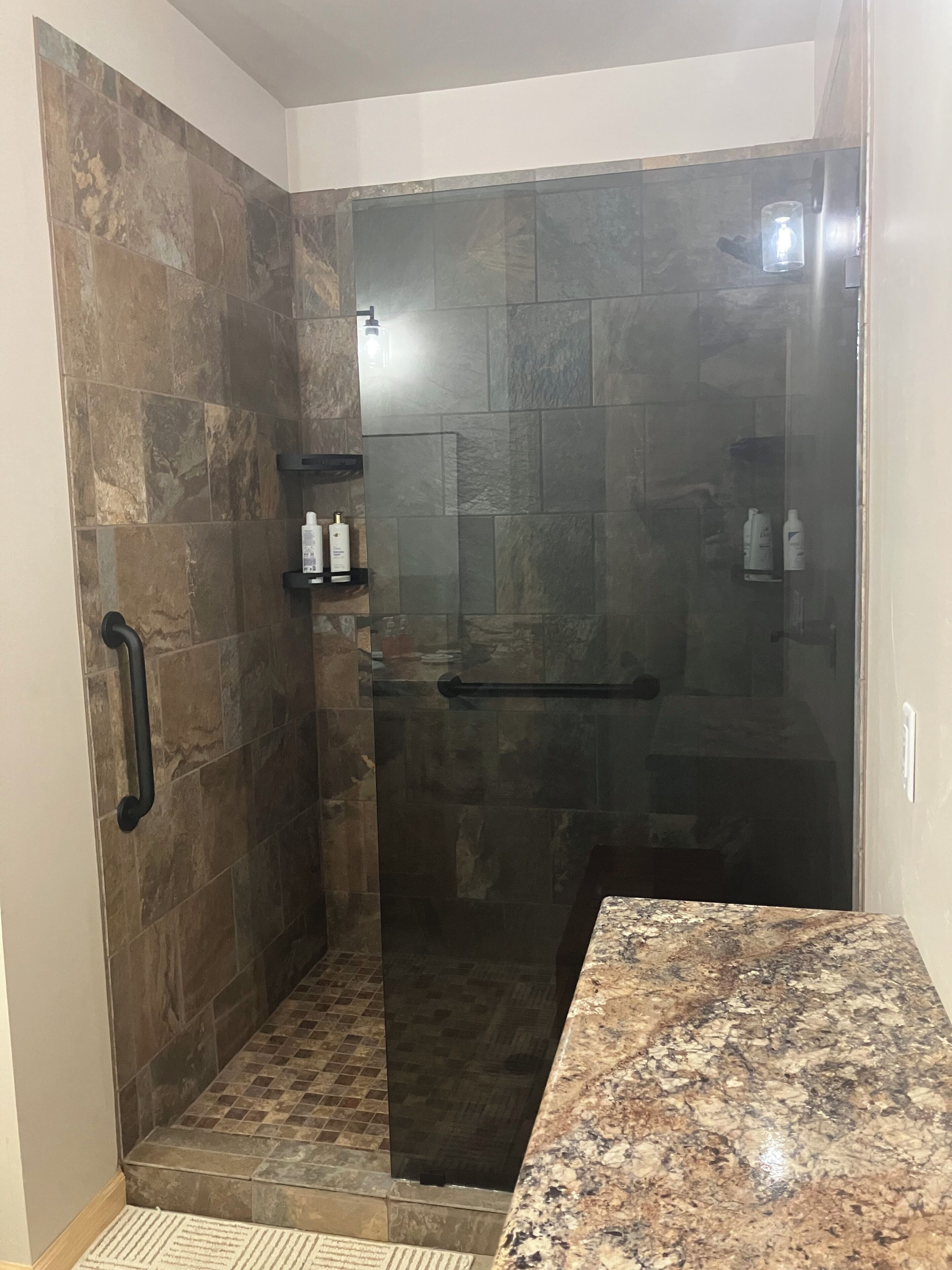 A shower area with a glass door, a handrail, and a shelf with toiletries.