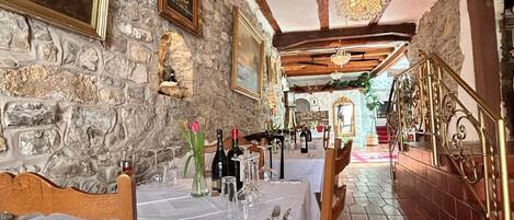 A dining area with stone walls, wooden beams, and a chandelier. Tables are set with white tablecloths and chairs.