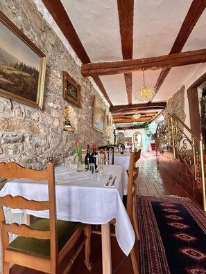 Une salle à manger aux murs de pierre, avec des poutres en bois et un lustre. Les tables sont dressées avec des nappes blanches et des chaises.
