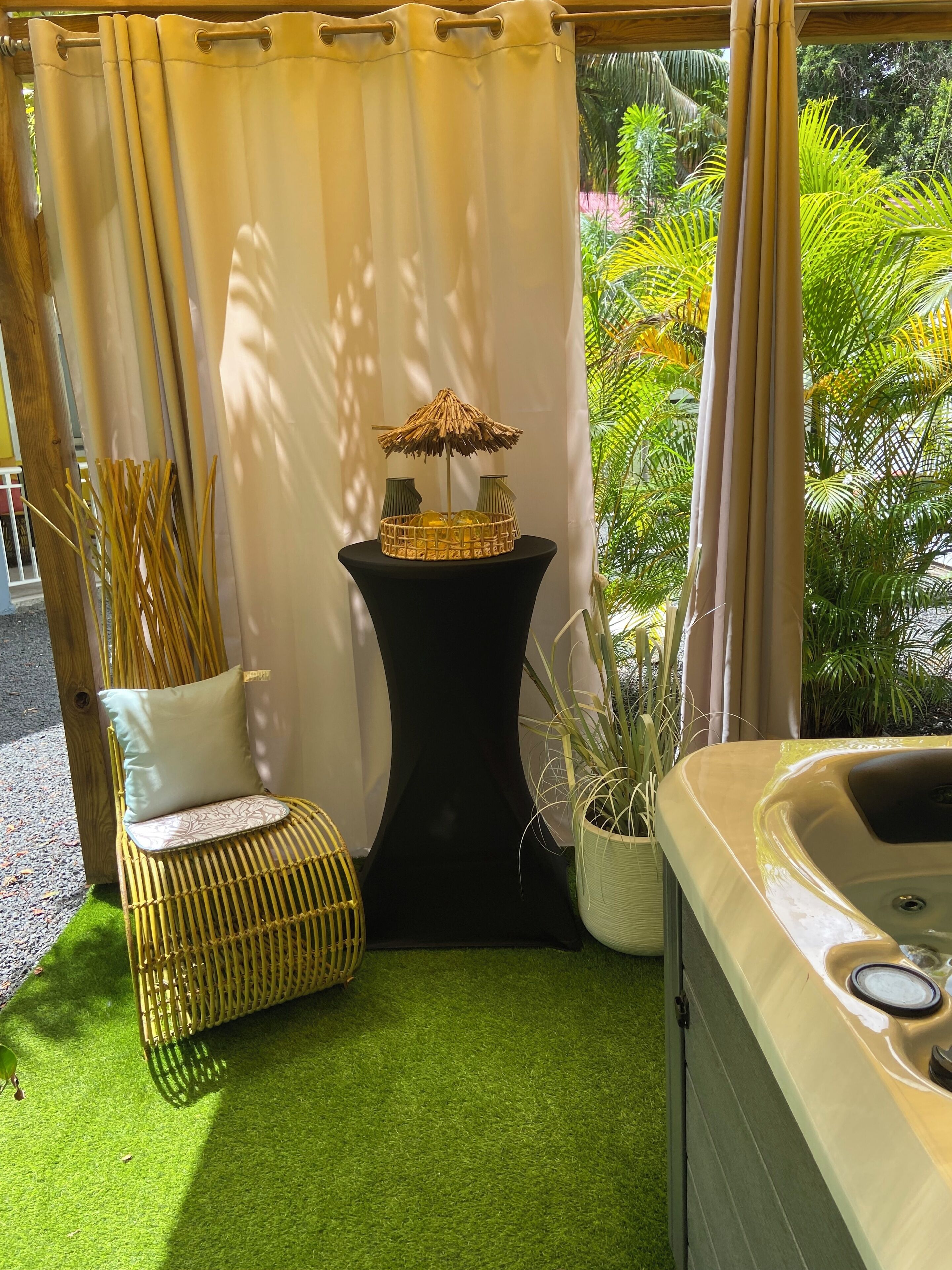 A room with a hot tub, a wicker chair with a cushion, a small table with a decorative item, and a curtain.