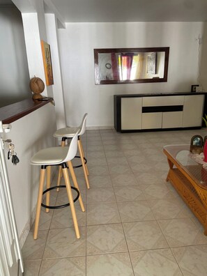 Une cuisine moderne avec un coin repas, comprenant un plan de travail blanc, deux chaises, un tabouret en bois et un meuble surmonté d’un miroir.