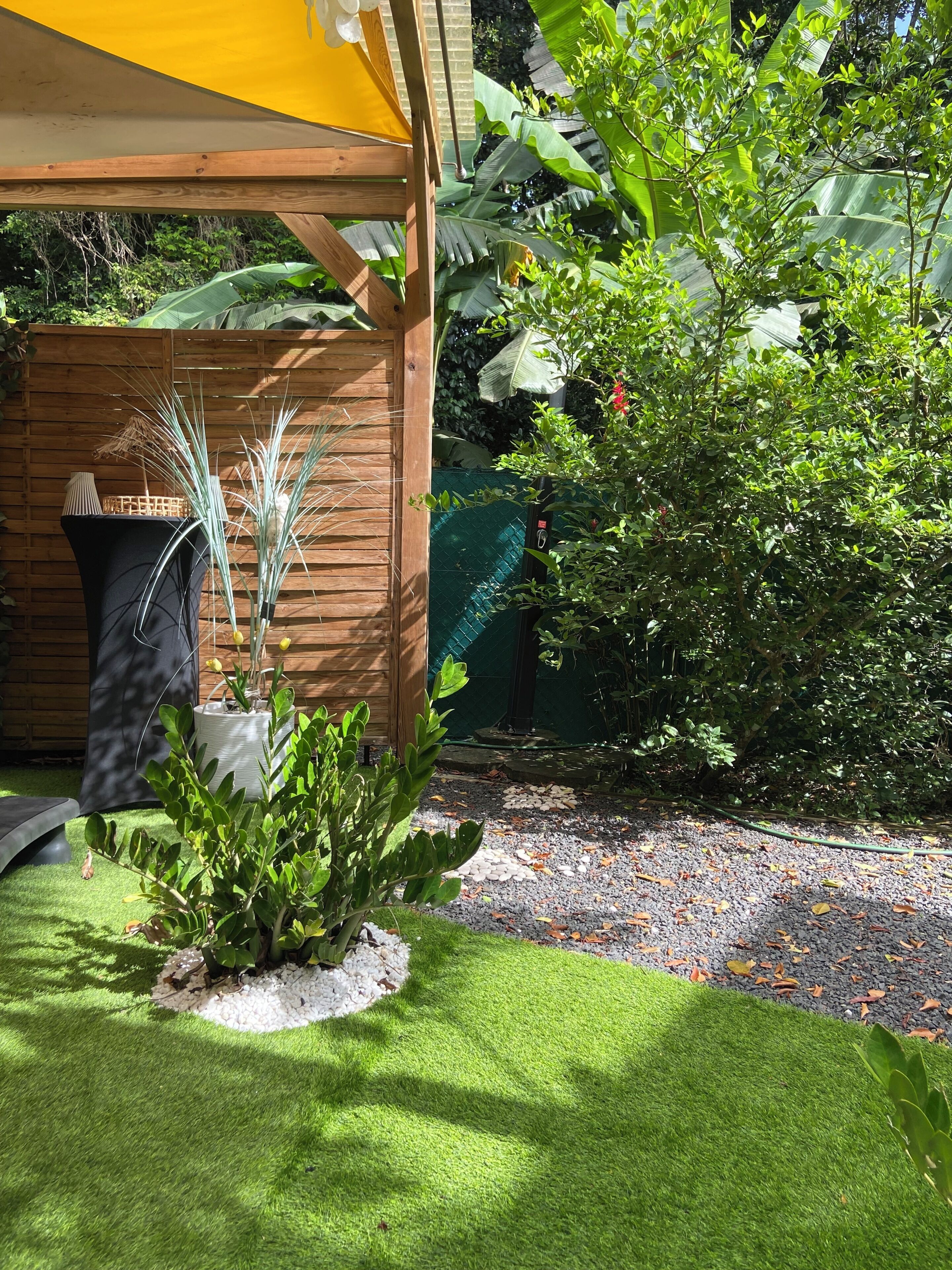 Área de jardim com pérgola de madeira, plantas em vasos e caminho de cascalho.