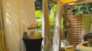 A covered outdoor seating area with a wicker chair, a small table with a vase, and a hot tub. - Mon petit coin de paradis - Spa privatif & calme tropical (Morne-à-l'Eau)