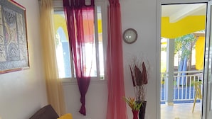 A living room with a brown sofa, yellow pillows, a glass coffee table, and a view of a yellow building through a window with red curtains. - Mon petit coin de paradis - Spa privatif & calme tropical (Morne-à-l'Eau)