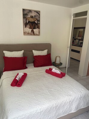 Une chambre d’hôtel avec un lit, deux oreillers rouges, une couvre-lit blanche, un tableau au mur et une table de chevet avec une lampe.
