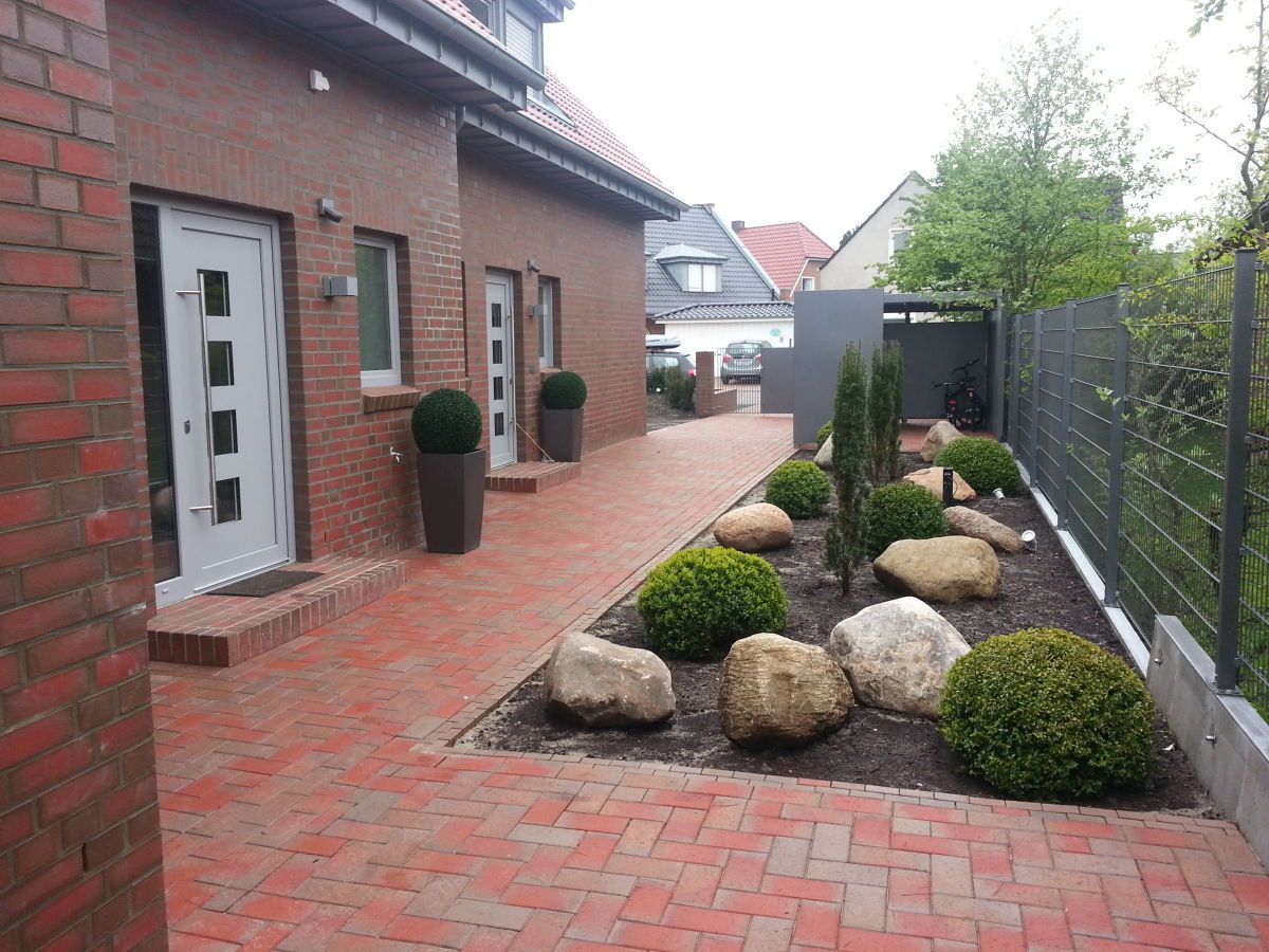 A brick pathway leading to a house with a white door, surrounded by neatly arranged rocks and shrubs.