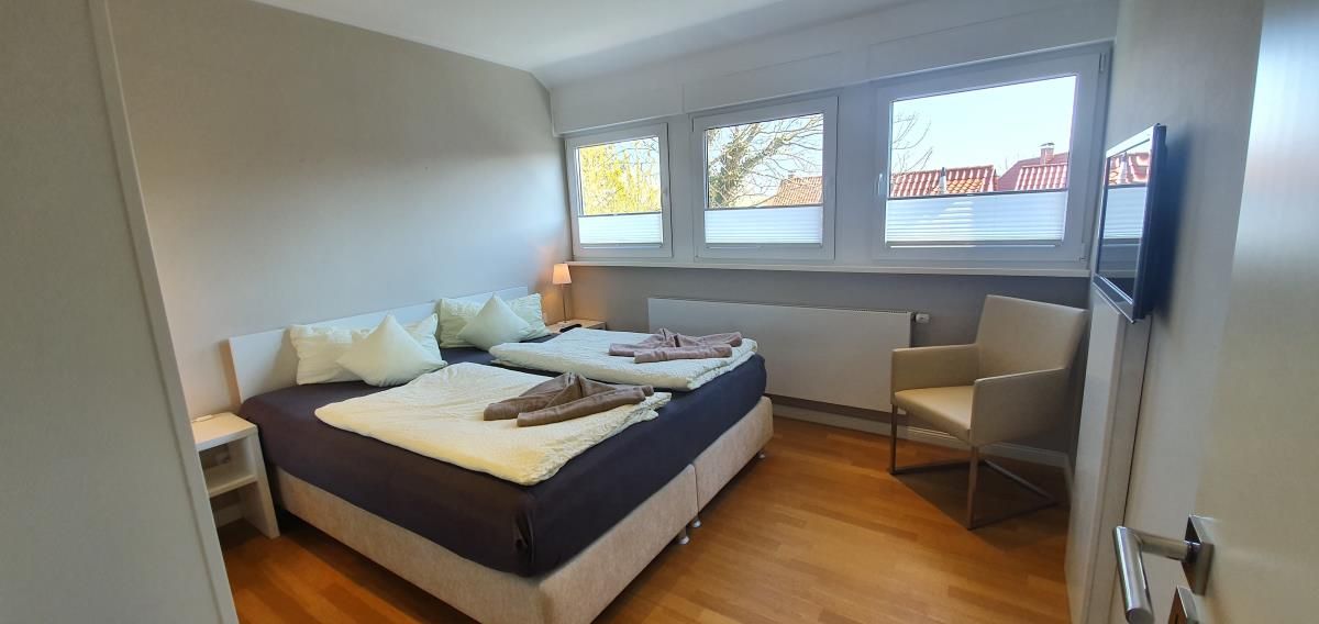 Quarto com duas camas, uma cadeira, televisão e janela com vista para edifícios.