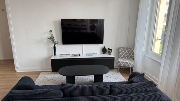 A modern living room with a large flat-screen TV mounted on a white wall, a black sofa, a coffee table, and a patterned armchair. - Nice Apartment in Cherbourg for 4 Guests (Cherbourg-en-Cotentin)