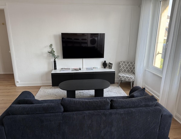 Una sala de estar moderna con un televisor de pantalla plana grande montado en una pared blanca, un sofá negro, una mesa de centro y un sillón estampado.