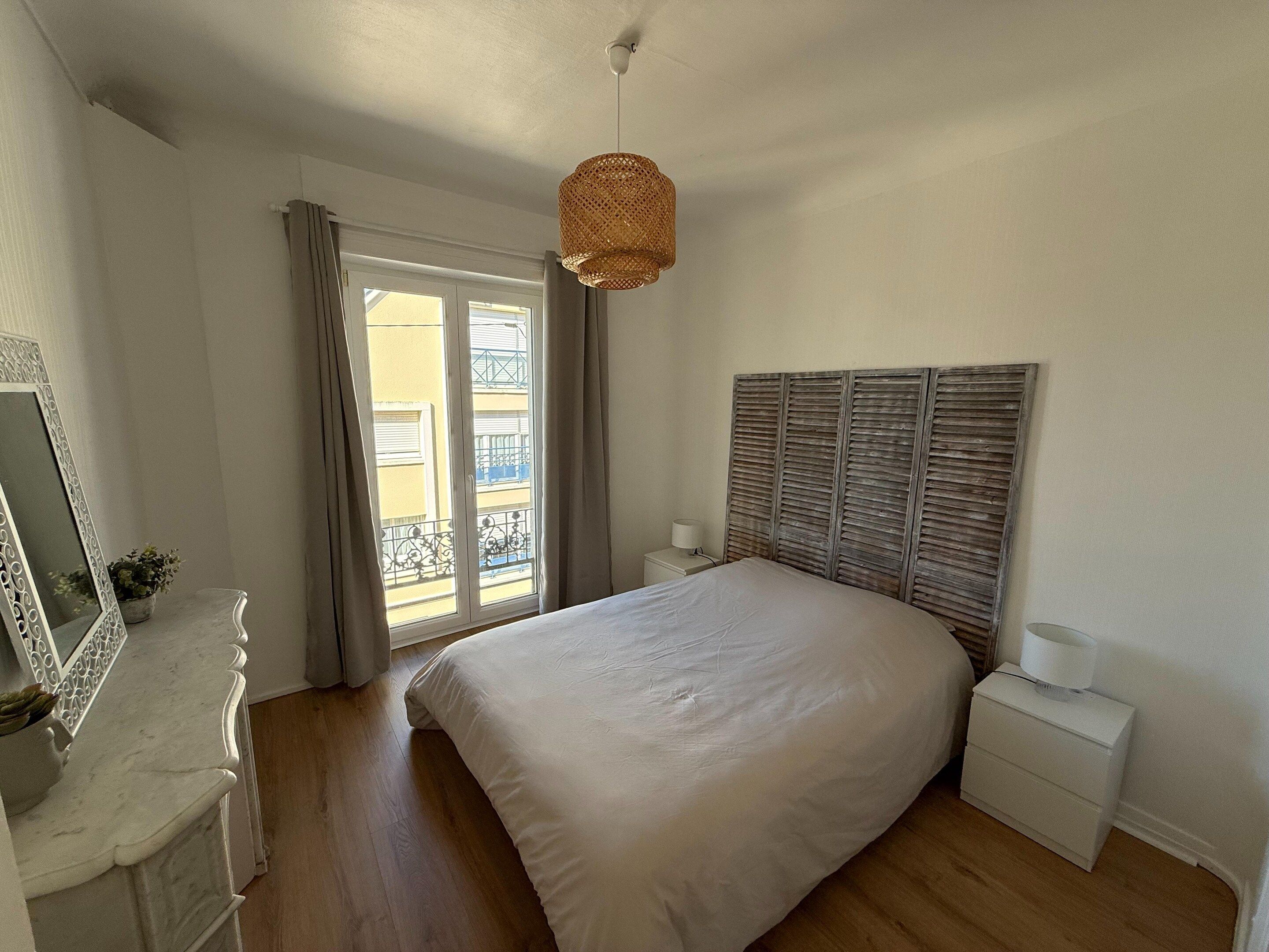 A bedroom with a large bed, a bedside table, a nightstand, a hanging light fixture, and a window with a view of buildings.