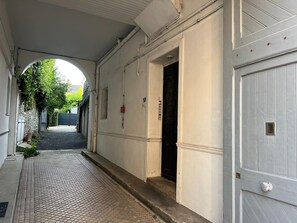 Un pasillo con piso de baldosas, una puerta con placa de número y un detector de incendios instalado en la pared.