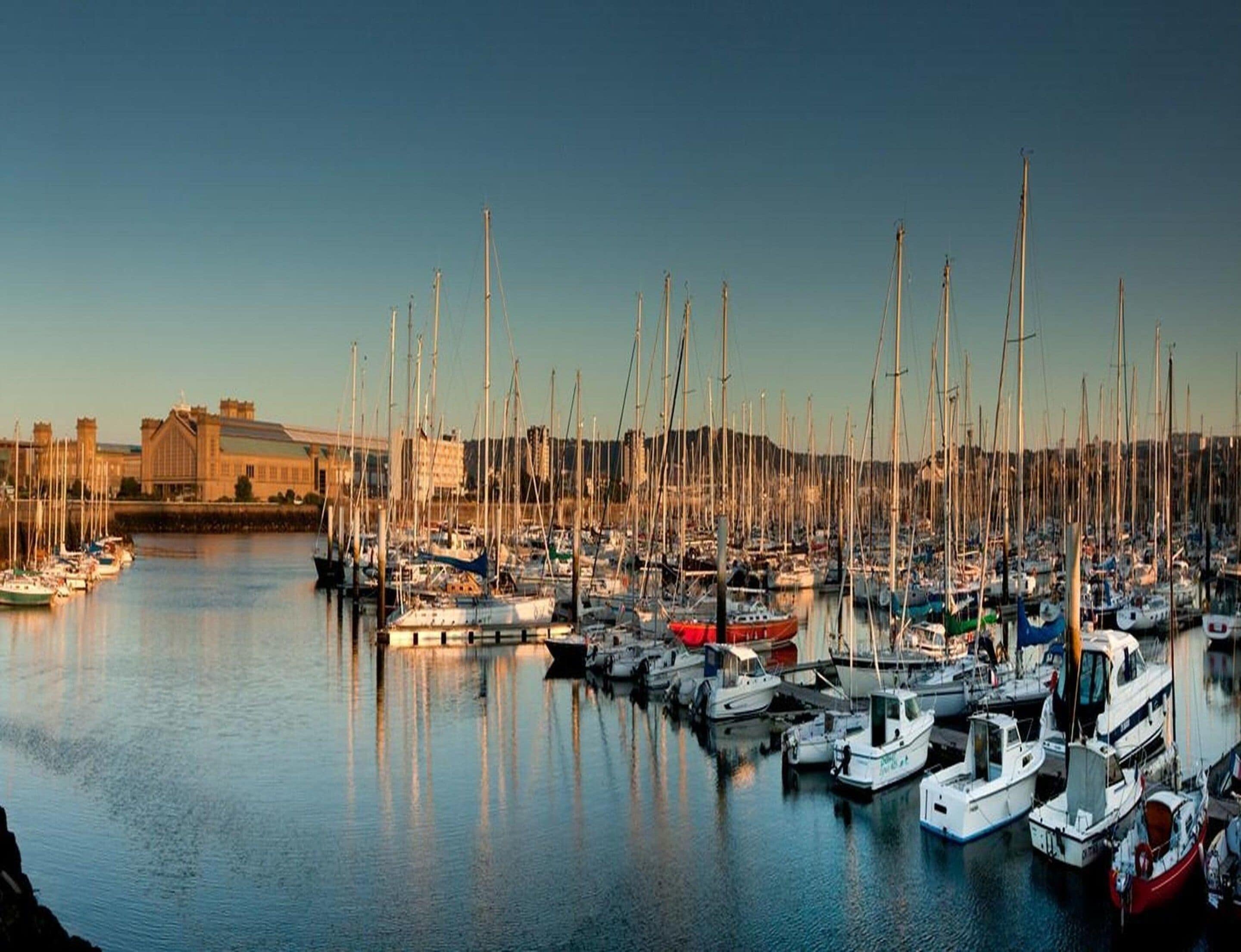 A marina with numerous boats docked, a building with a large roof, and calm water reflecting the boats.