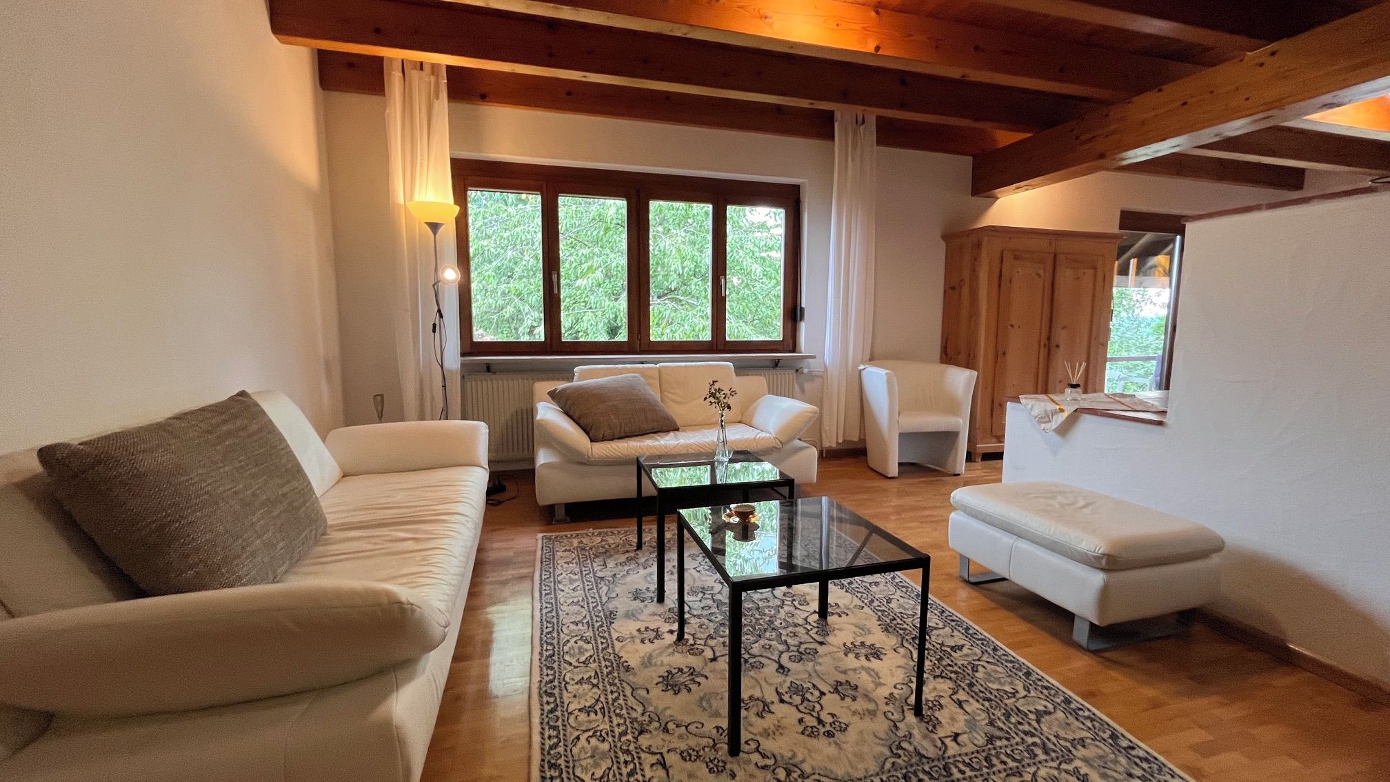A living room with a sofa, a coffee table, and a chair. There is a window with curtains and a view of greenery outside.