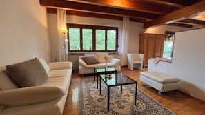 A living room with a sofa, a coffee table, and a chair. There is a window with curtains and a view of greenery outside. - Koppelblick Vacation Home - My SWH (Murg)