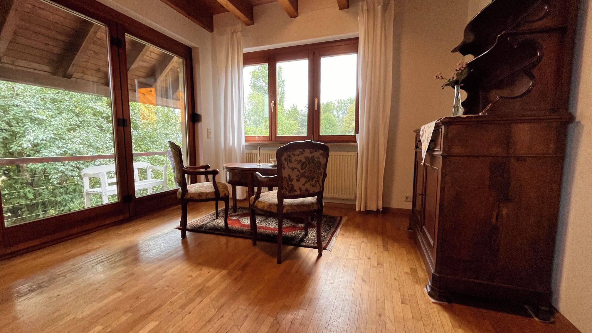 Ein Zimmer mit Holzboden, einem Esstisch mit Stühlen, einem Heizkörper und Blick auf Grünflächen durch große Fenster.
