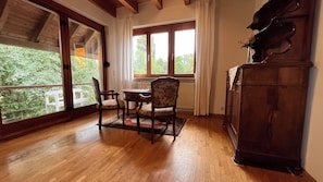 A room with a wooden floor, a dining table with chairs, a radiator, and a view of greenery outside through large windows.