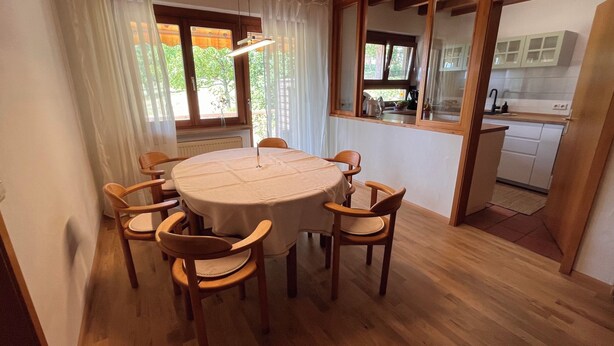 Un comedor con una mesa redonda y seis sillas, una ventana con cortinas y vistas a una cocina.