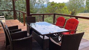 Terraza exterior con mesa de vidrio y sillas de mimbre, rodeada por barandilla de madera y follaje.